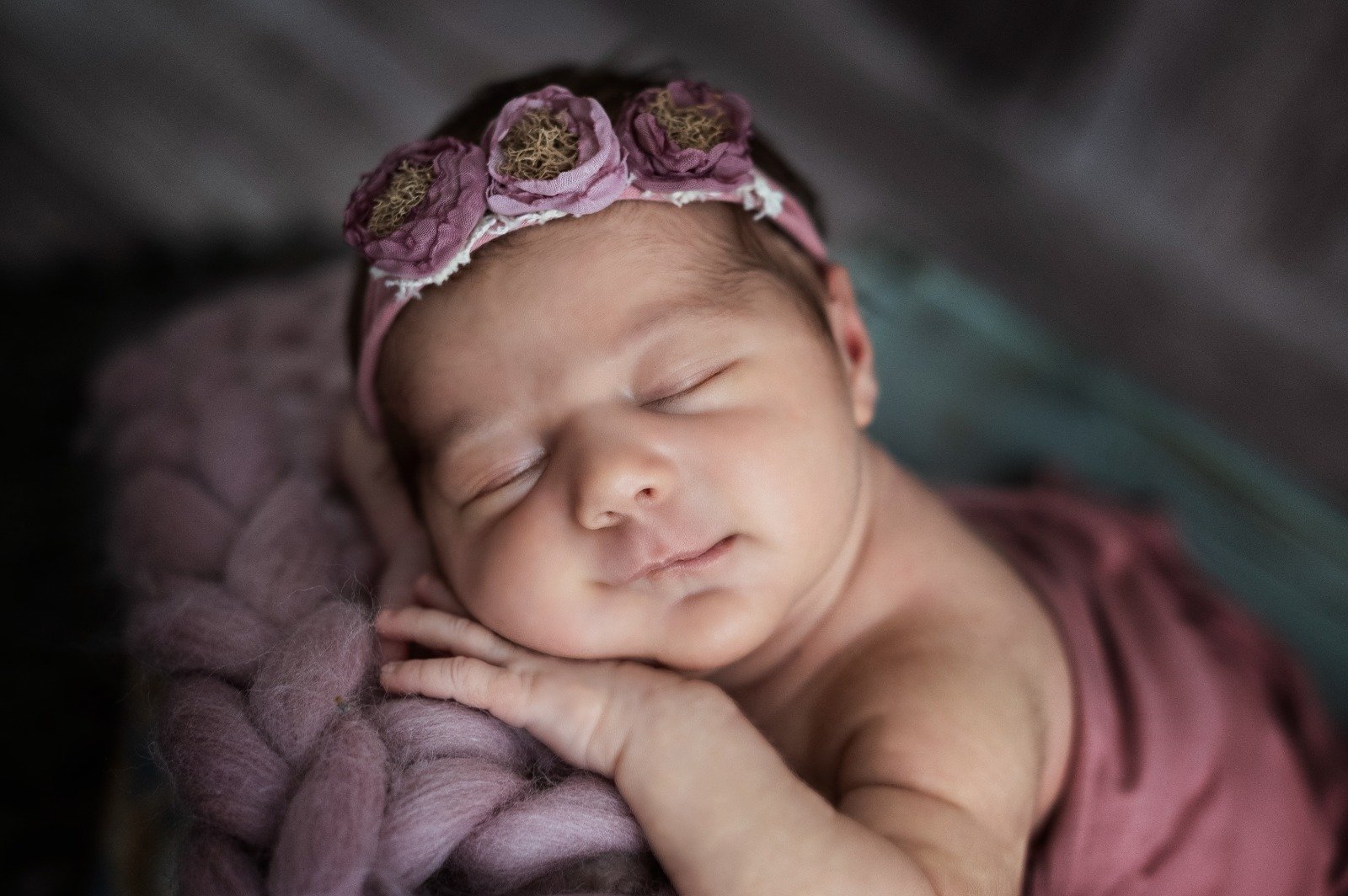 Neonato addormentato con fascia per capelli rosa decorata con fiori viola, poggiato su una coperta morbida.